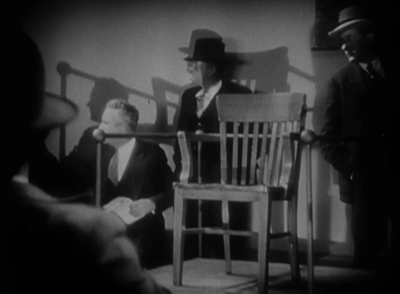 Clear black-and-white frame showing a wooden chair on a raised platform, men in hats around it and strong expressionist shadows on the wall.