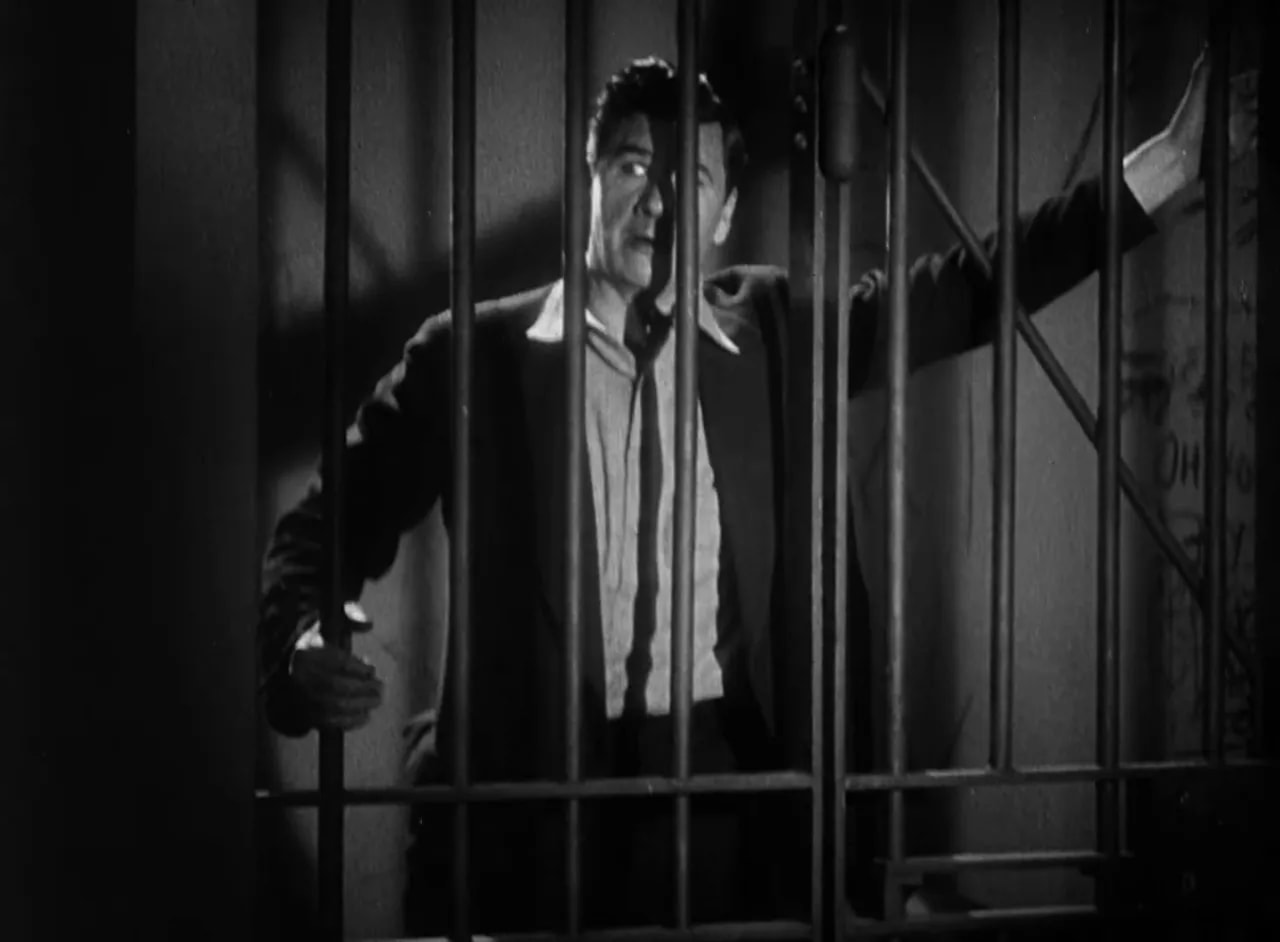 Protagonist behind prison bars with clear chiaroscuro lighting, face visible between bars.