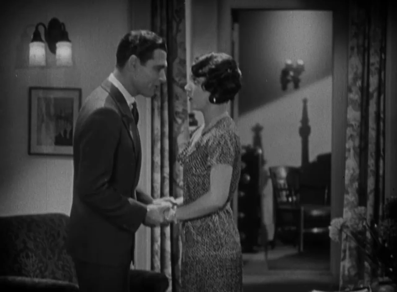 Clear black-and-white still of a man and woman holding hands in a living room, showing expressive body language
