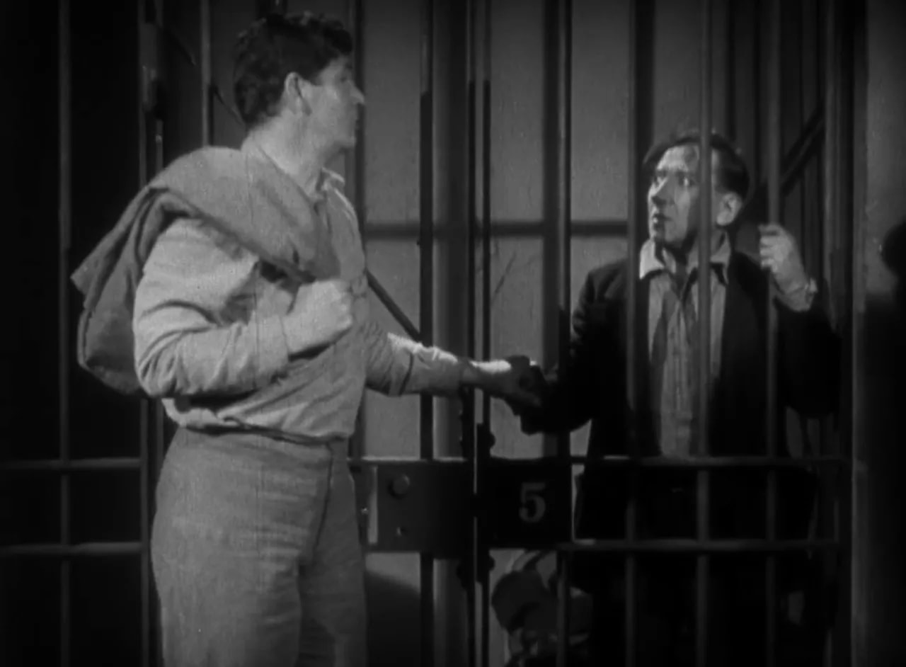 Two men at a cell door: a man outside with a jacket over his shoulder and a prisoner holding the bars inside.