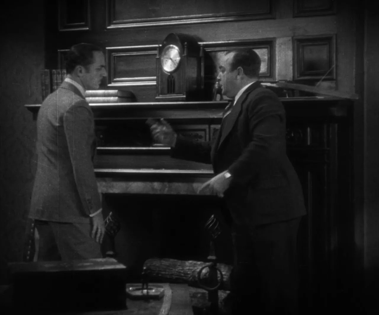 Two men at a mantelpiece with books and a clock as one explains a concealed mechanism in a black-and-white frame
