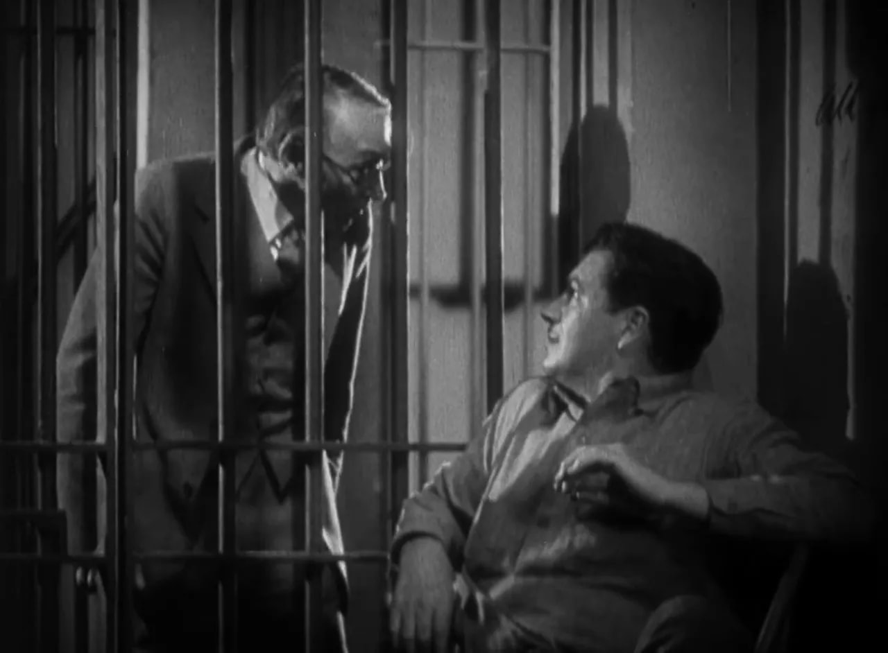 Warden leaning into a cell to speak with a seated inmate; clear, high-quality black-and-white still
