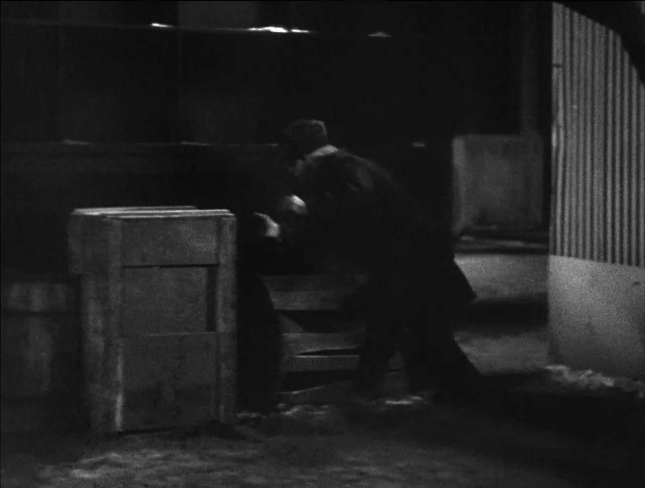 Black-and-white scene of a man crouching or hiding beside crates and walls at night